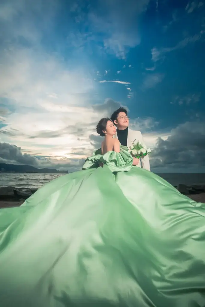 Bride in flowing green gown holding bouquet by the sea in Penang under moody sky—captured by wedding photographer Struck By Luna.