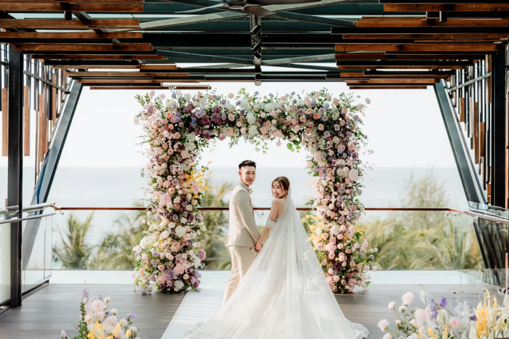 Bride and groom standing under a colorful floral arch on a balcony overlooking the ocean during a romantic outdoor wedding ceremony by Struck By Luna