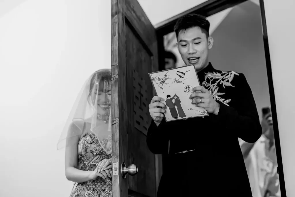 Bride in Chinese wedding dress behind door as groom holds illustrated vow booklet—captured in Penang by wedding photographer Struck By Luna.